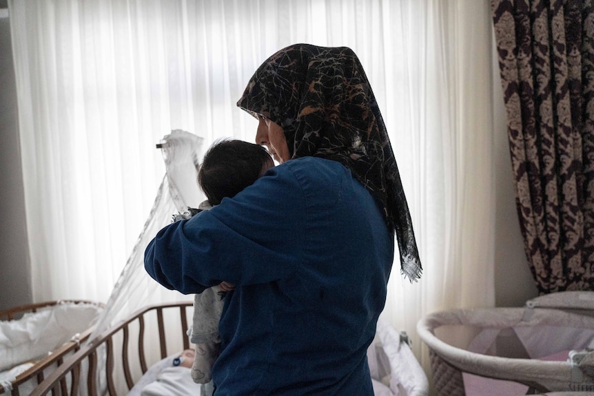 A Turkish nanny wearing a hijab holds a tiny baby in a small room.