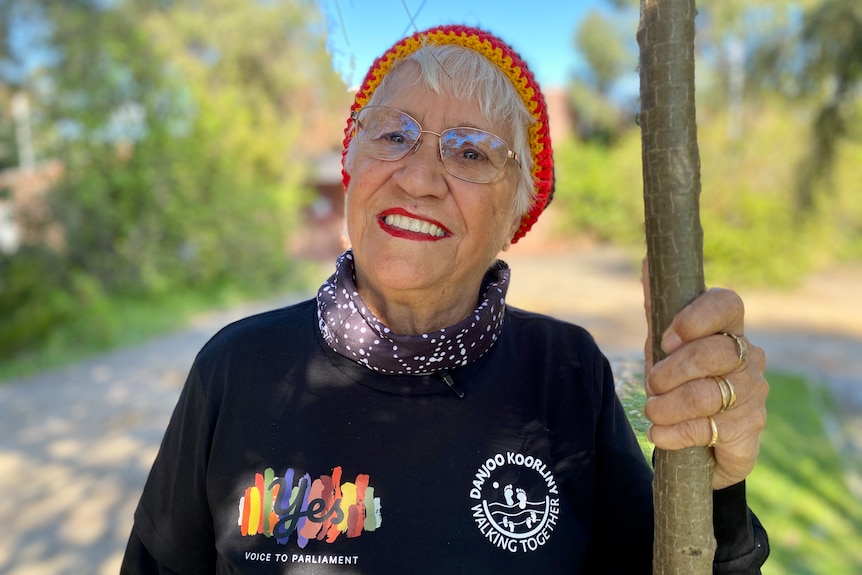 Elderly Aboriginal woman wearing beanie and glasses holding on to tree smiling at camera.