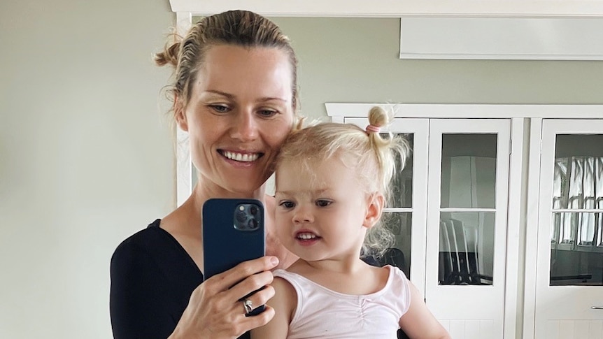 Blonde-haired woman holding a girl toddler and phone takes a selfie in the mirror of an empty room with polished wood floors.