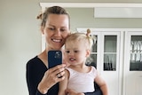 Blonde-haired woman holding a girl toddler and phone takes a selfie in the mirror of an empty room with polished wood floors.