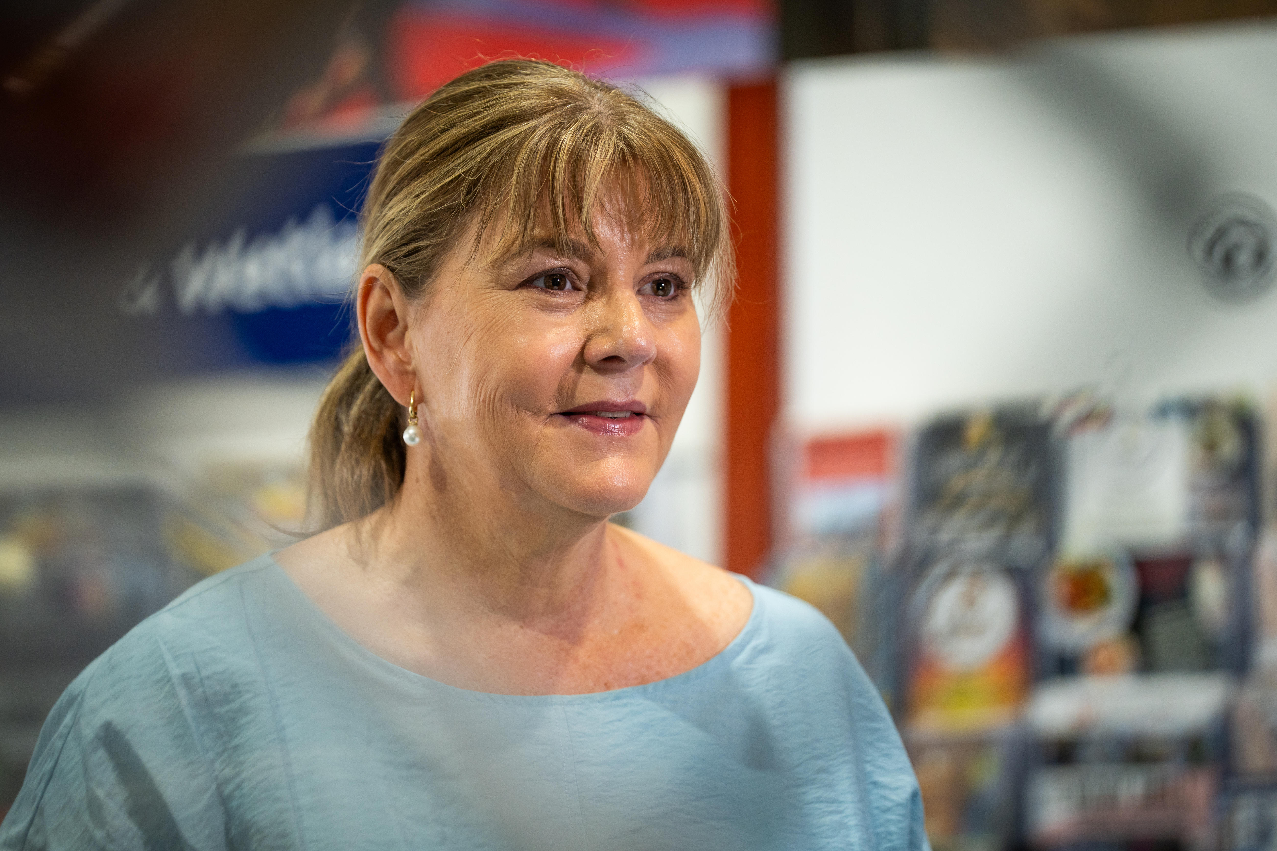 Mujer blanca mirando a la cámara a la derecha, cabello castaño recogido en cola de caballo, flequillo, blusa azul claro, tienda borrosa en el fondo.