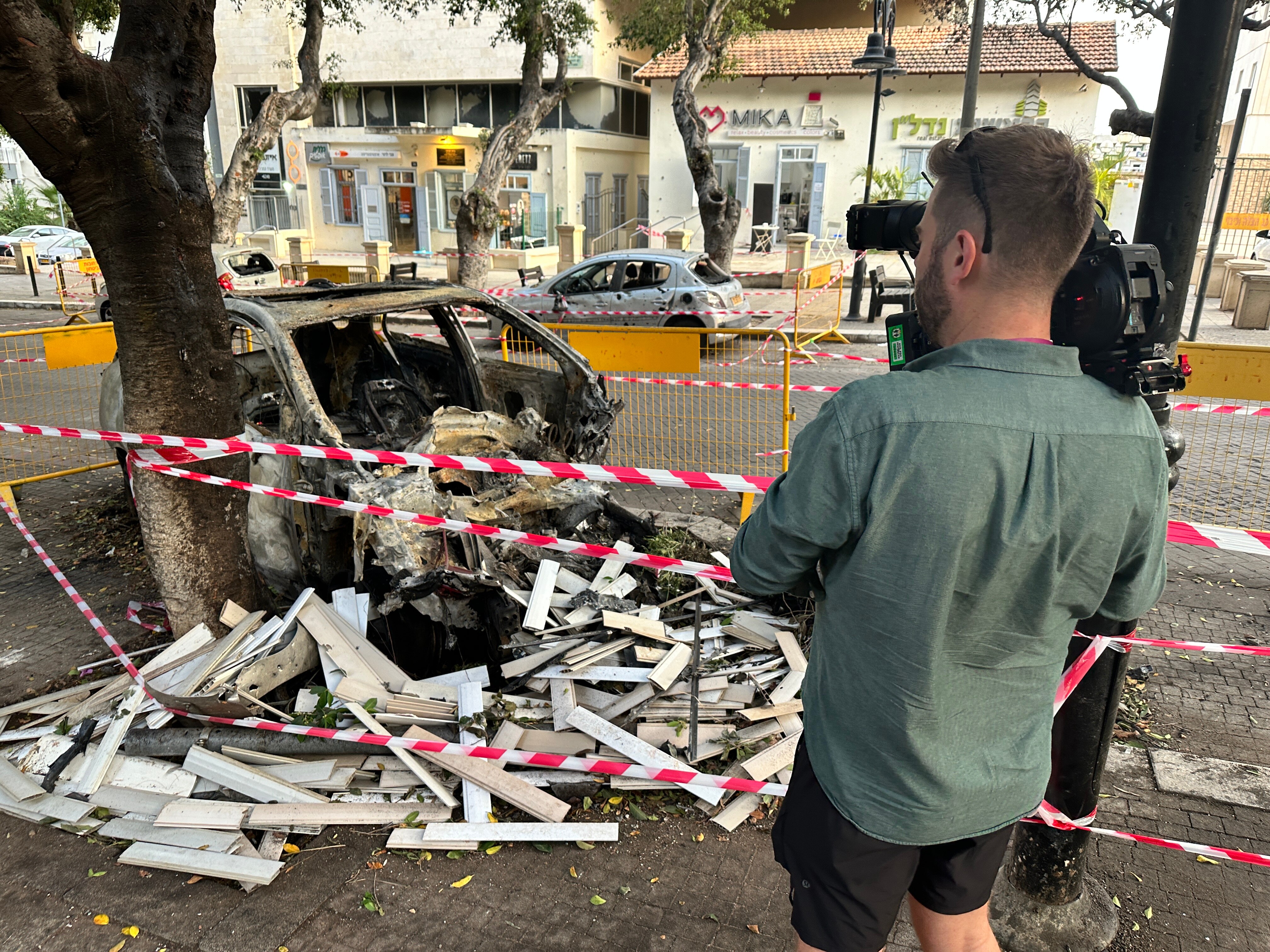 Cameraman filming burnt out car in a street, surrounded by police tape.