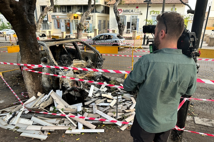 Cameraman filming burnt out car in a street, surrounded by police tape.