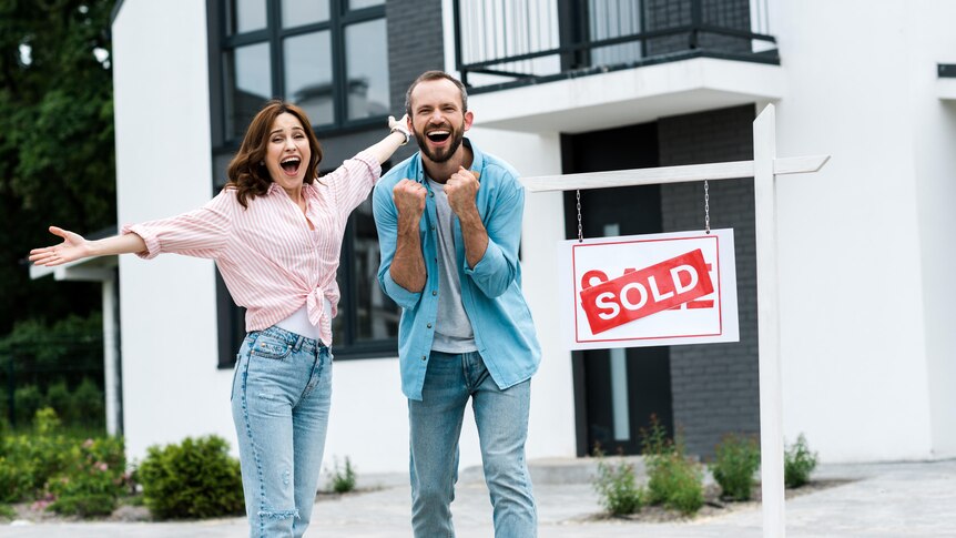 A woman and a man look excited in front of a new townhouse with a 'sold' sign out the front.