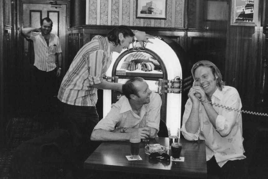Four men sit around a juke box laughing, with one speaking on a phone