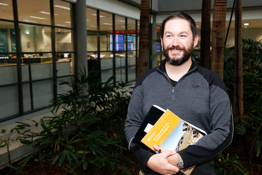 Man with a beard smiles as he holds a book on the history of earthquakes in Canberra.