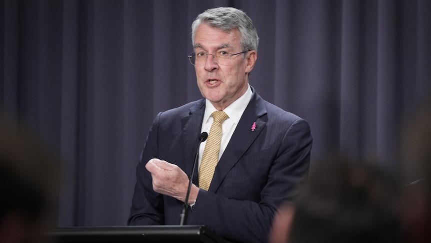 A medium shot of a man in a suit and tie standing and speaking into a microphone at a lecturn. He holds up one clenched hand