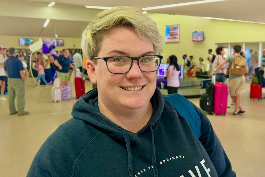 A woman with short blonde hair, wearing glasses and a hoodie, standing in an airport.