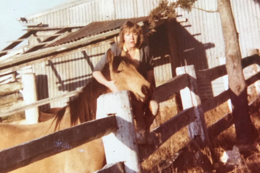 Tracey Higgins, aged in her 20s, sits on a paddock fence with her arm around a horse next to her. 
