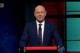 A video still of Tom Gleeson on the set of Hard Quiz standing behind the podium looking at the camera
