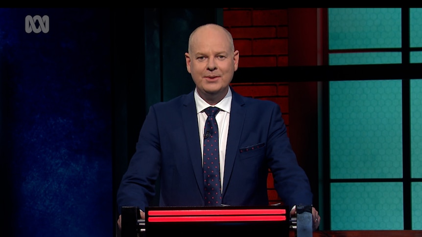 A video still of Tom Gleeson on the set of Hard Quiz standing behind the podium looking at the camera