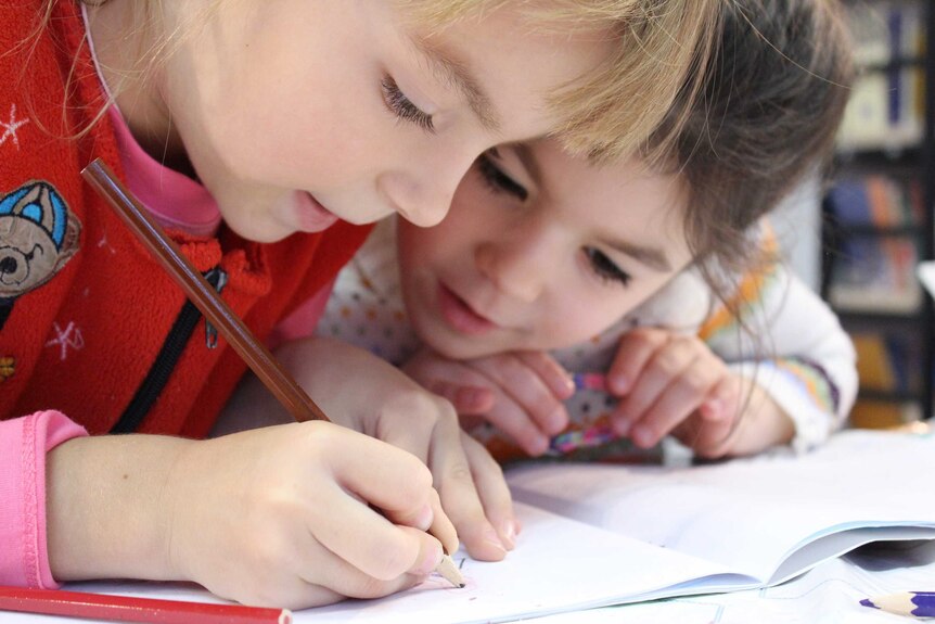 Two young girls at a table lean over book where one of the little girls draws with a brown pencil