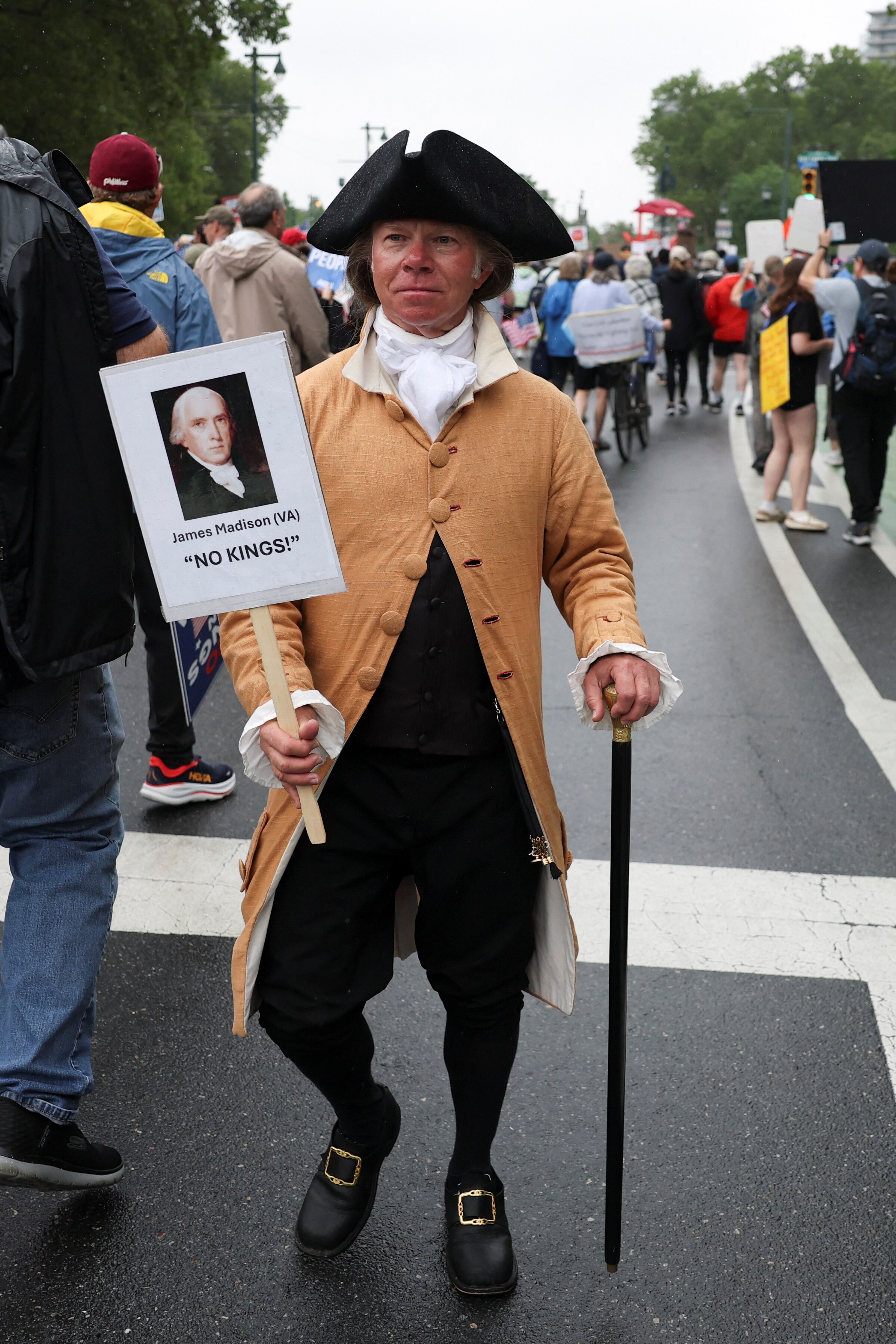 A man wearing 18th century garb holds up a placard with a portrait of James Madison that reads "NO KINGS"