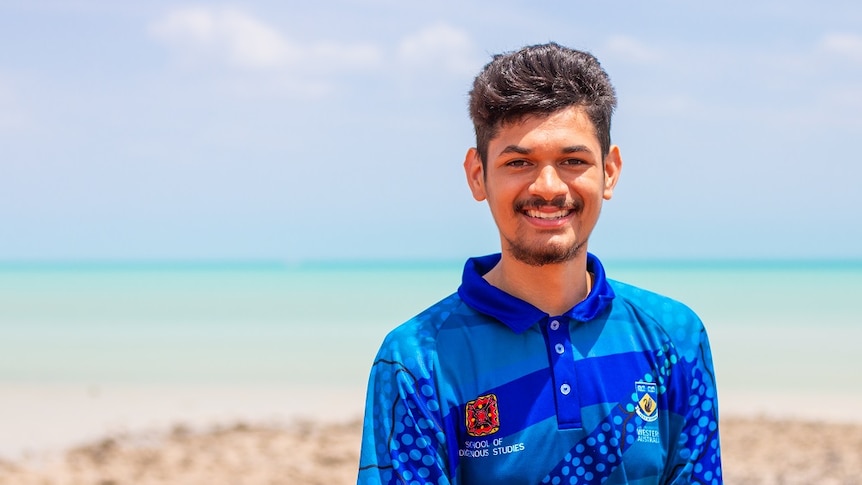 Young indigenous man with blue shirt stands on beach in front of turquoise water