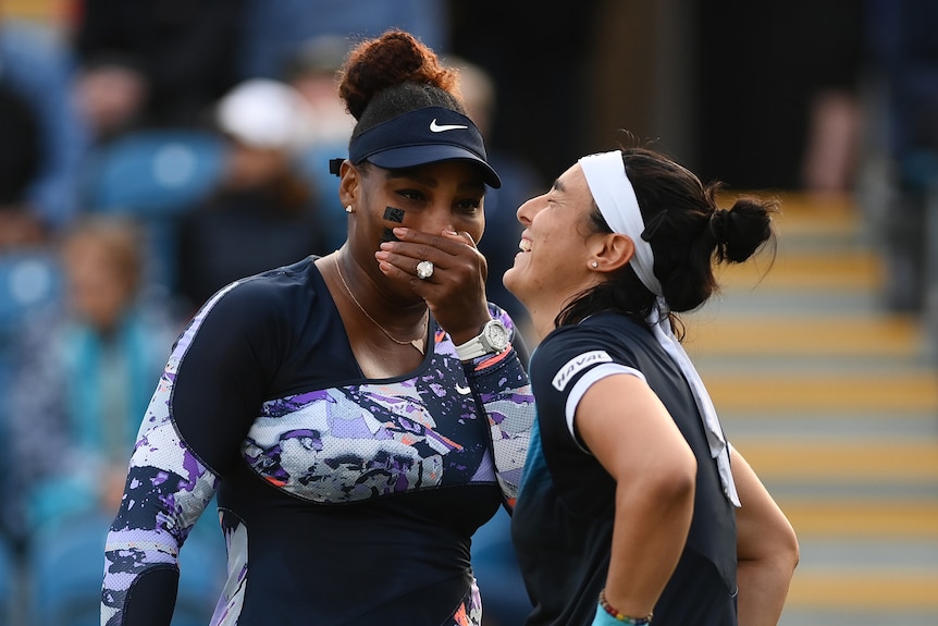 Serena Williams talks behind her hand as doubles partner Ons Jabeur throws her head back and laughs on court during a match.