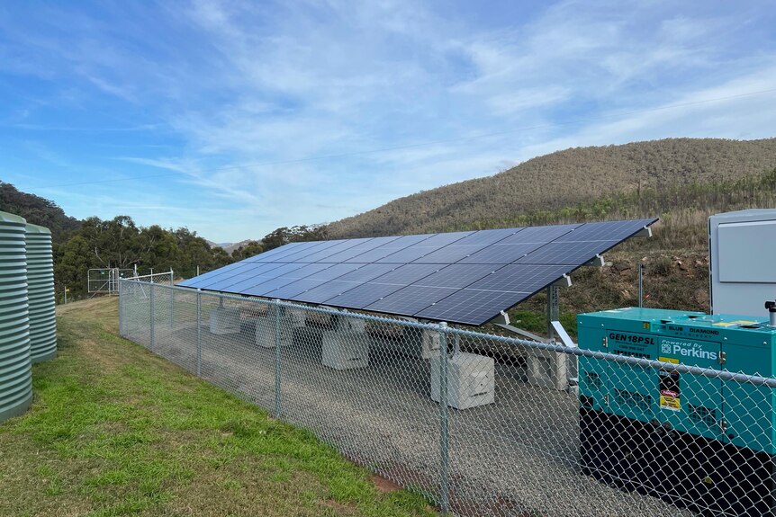 A set of solar panels and a generator behind a fence on a rural property.