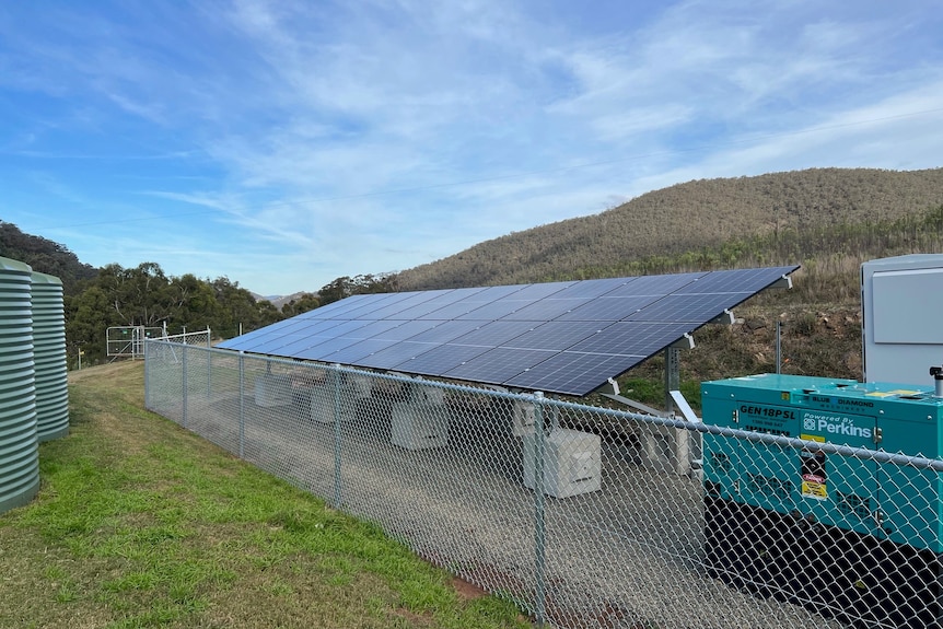 A set of solar panels and a generator behind a fence on a rural property.