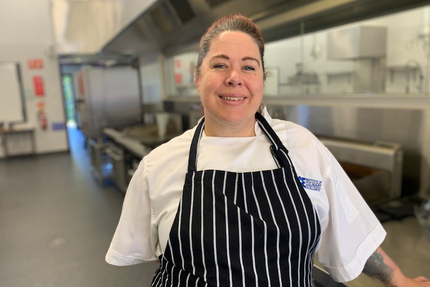 Image of a female chef smiling in a kitchen. She has her hair tied back and is wearing a striped apron over her whites