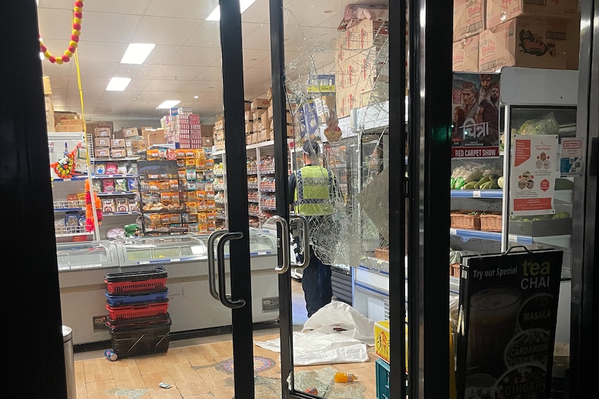 A police officer stands inside a Nepalese grocer following a vandalism incident, with a shattered glass door