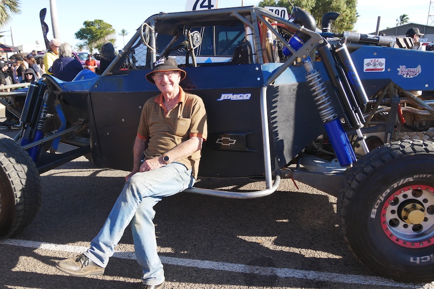 An older man sits on the running board of a racing buggy, smiling.