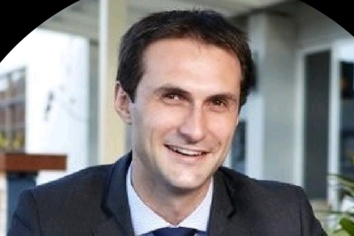 A young man in a dark suit smiles at the camera in a softly lit courtyard.