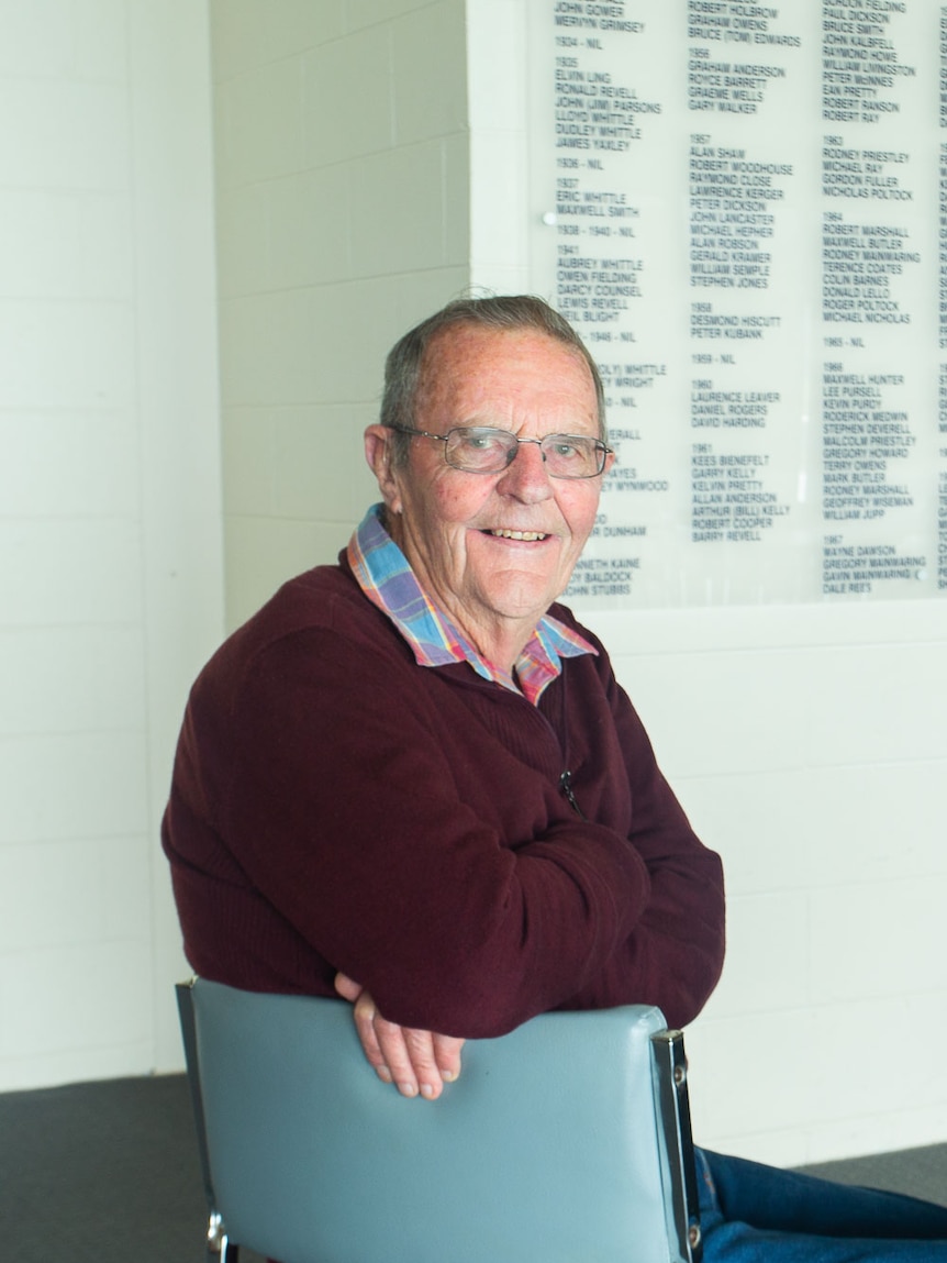 seated man smiles to camera inside surf club, honours board behind