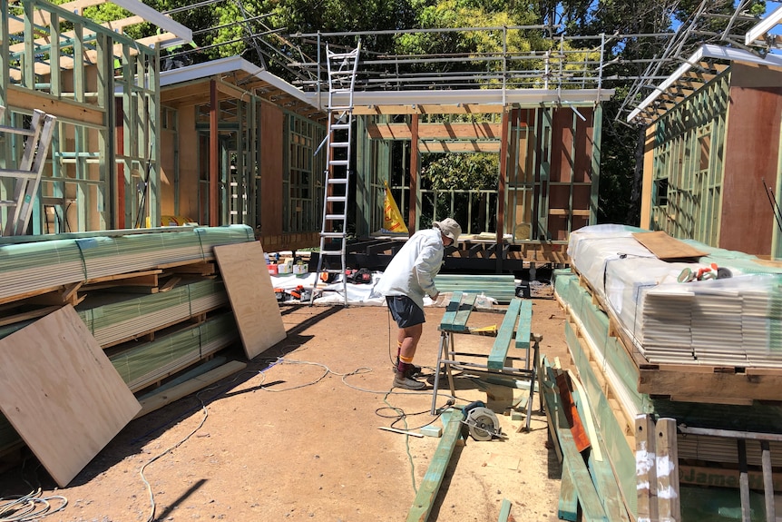 Builder measures timber on site surrounded by building supplies with three timber frames visible in background