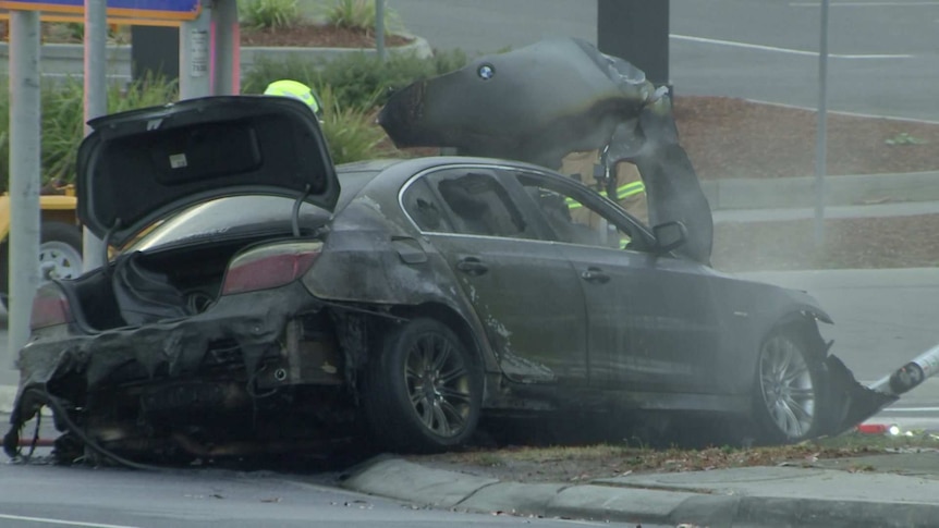 A burnt-out dark BMW car on the side of a road, with smoke coming from the bonnet.