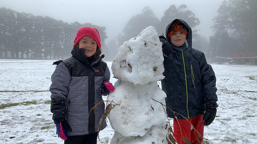 Two children in snow gear smile happily next to a snowman they have made in a field.