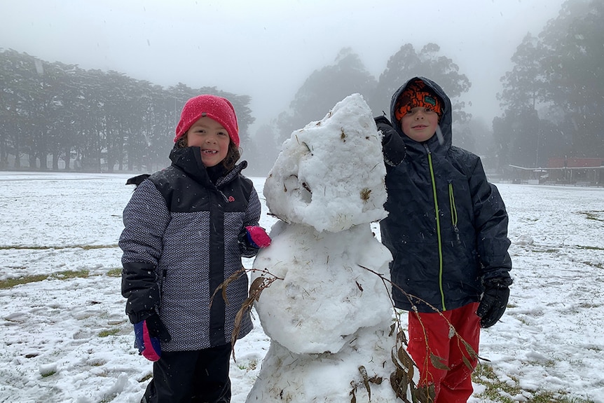 Two children in snow gear smile happily next to a snowman they have made in a field.