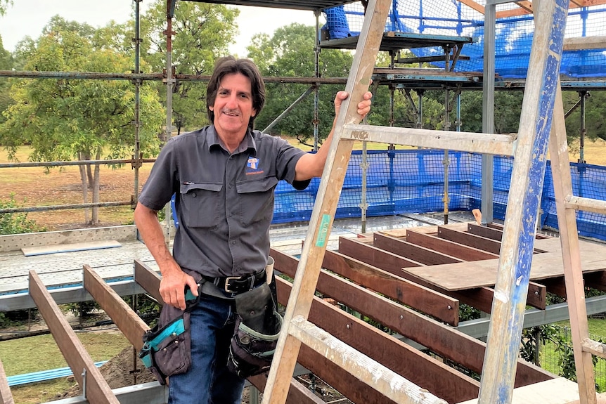 a man standing with a ladder on a builder site, wearing a tool belt.