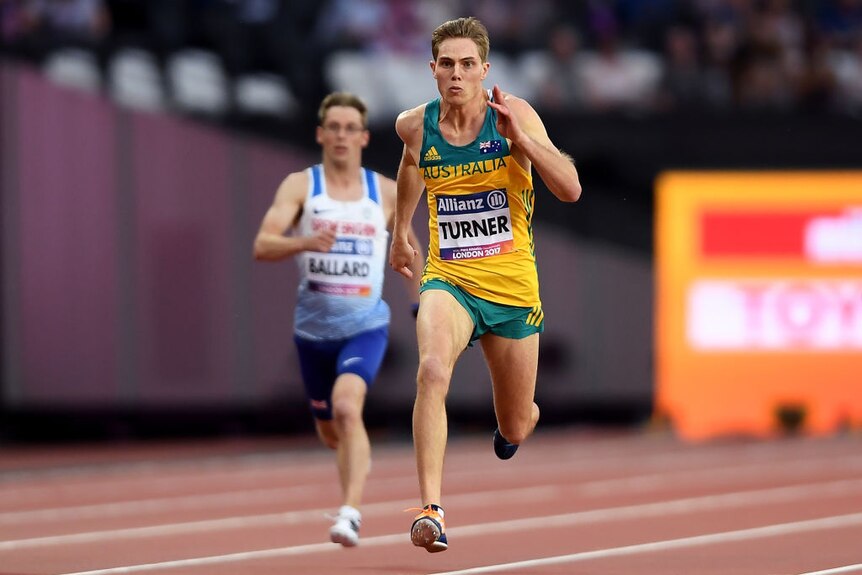 Blonde male in yellow top and green shorts running on Olympic track with runners behind him