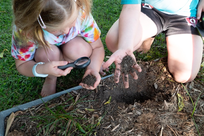 A young girl and boy crouch on a nature strip, holding soil in their hands.