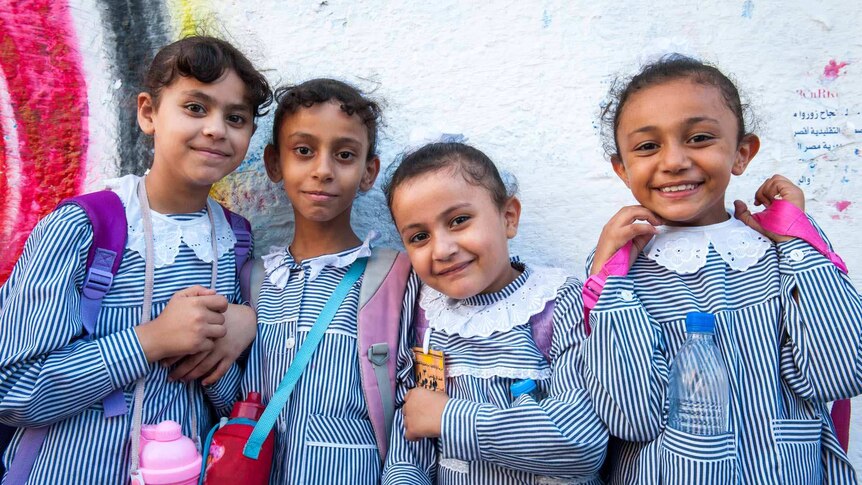 Four smiling primary school girls standing against a wall. They have blue-striped school uniforms and pink or purple backpacks.