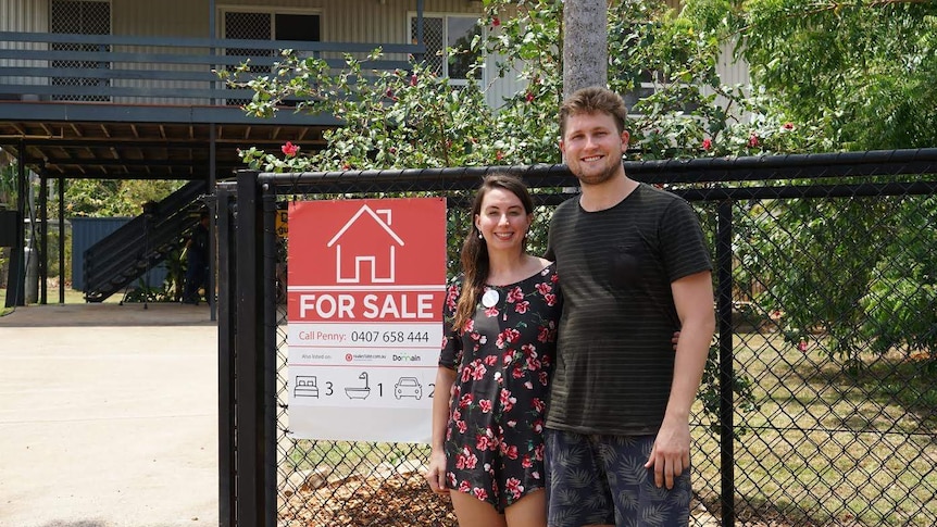 The pair stand outside the home, next to a "for sale" sign.