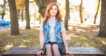 A woman with red hair sits on a bench with trees in the background.