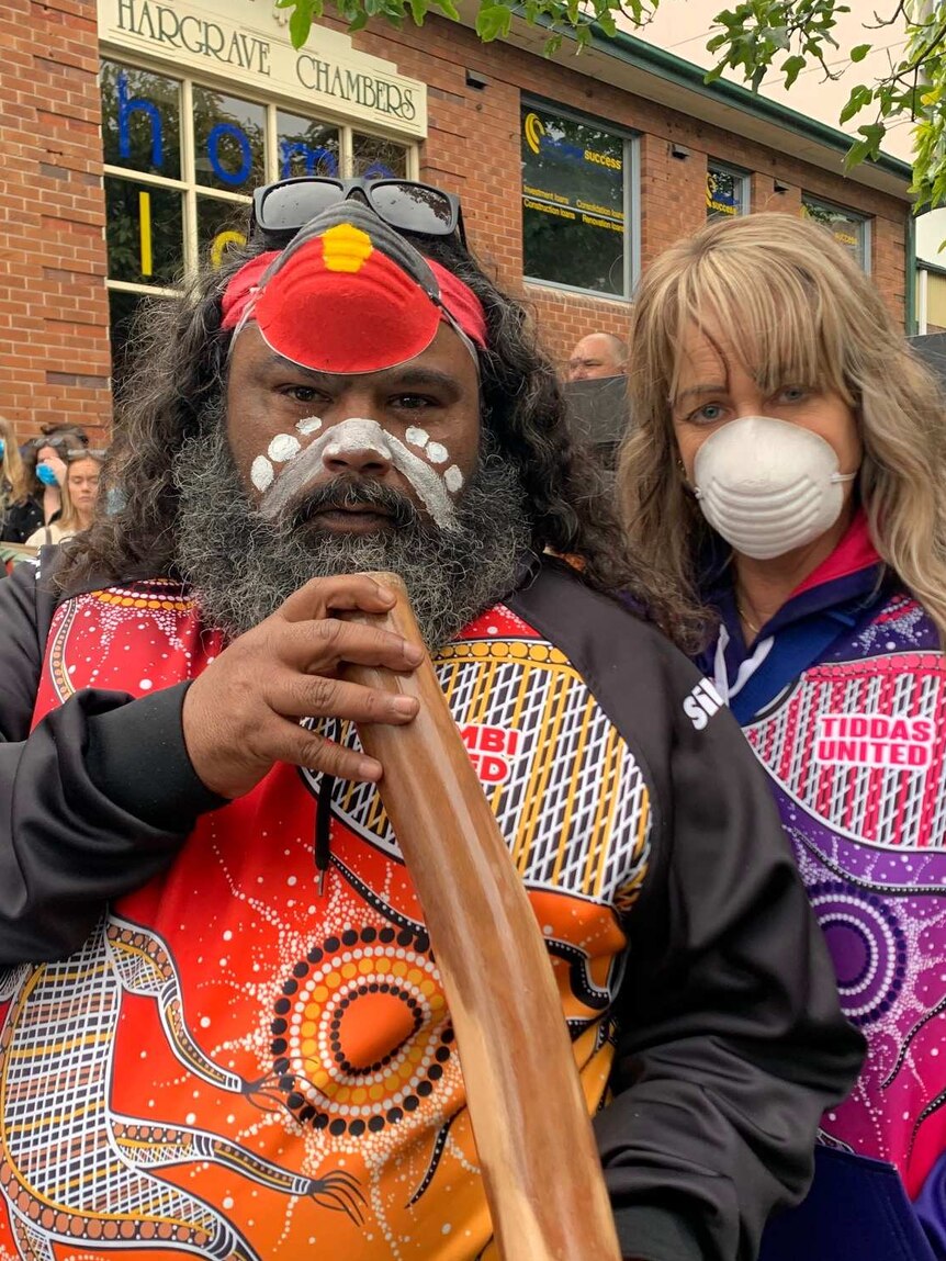 An Aboriginal man holds a didgeridoo with a woman standing beside him