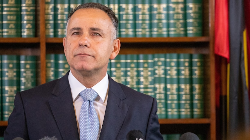 A man in a navy suit with a white shirt and light blue tie stands in front of a bookshelf.