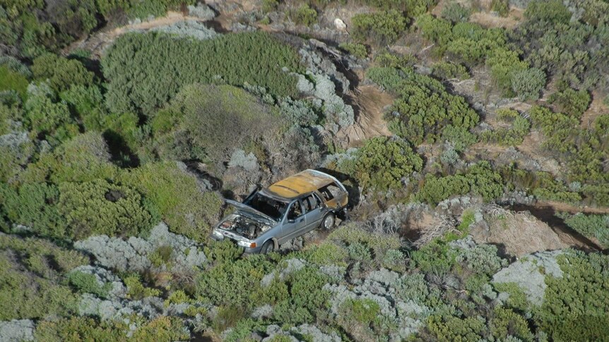 A burnt-out, older model station wagon in rugged bushland, as seen from above.