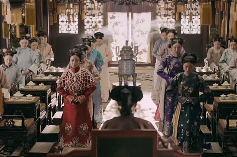 Four rows of Chinese women dressed in ancient Chinese costumes face a woman at the front of the room with her back to the camera