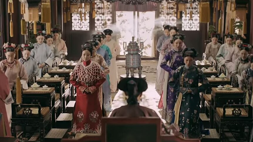 Four rows of Chinese women dressed in ancient Chinese costumes face a woman at the front of the room with her back to the camera
