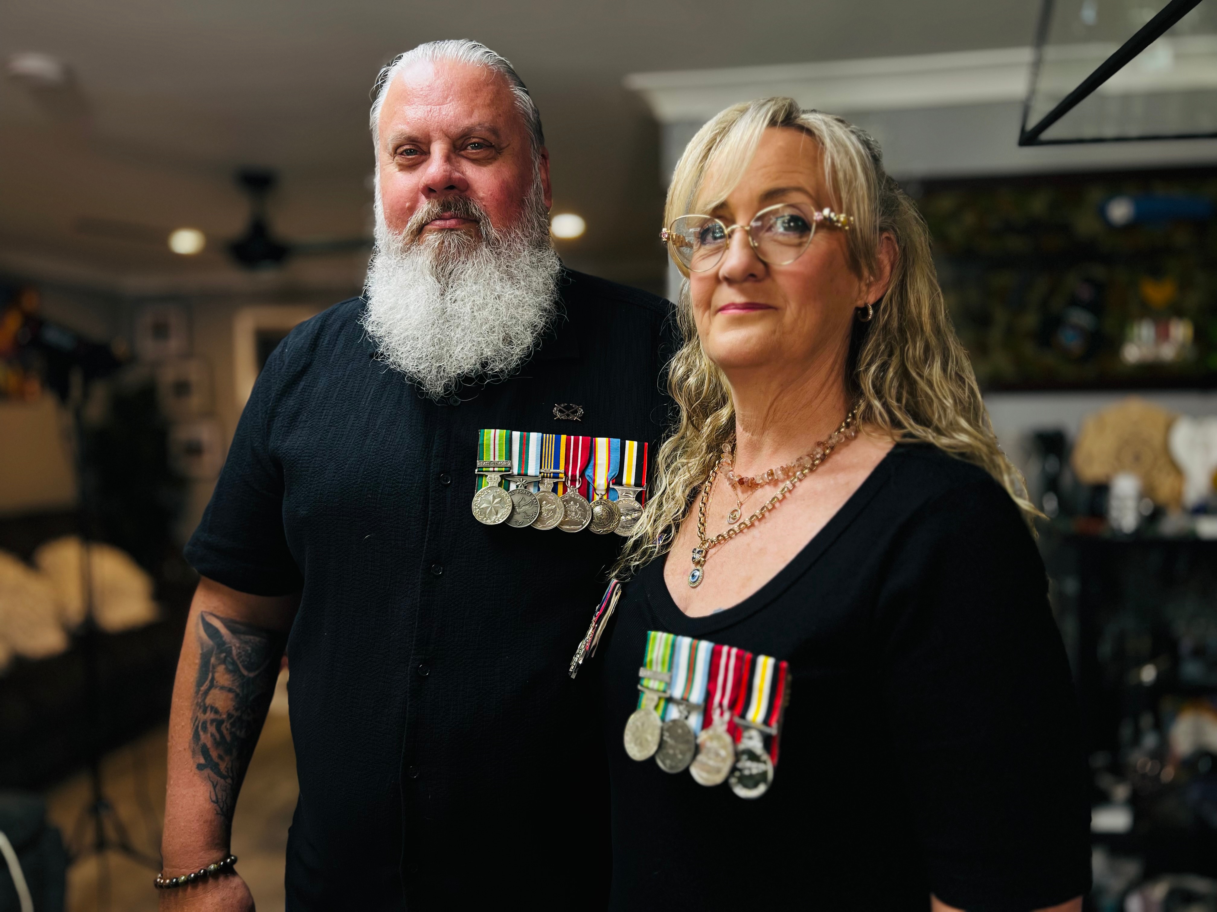 Two people stand inside a home wearing black but with several military medals on their chests. They look at the camera.