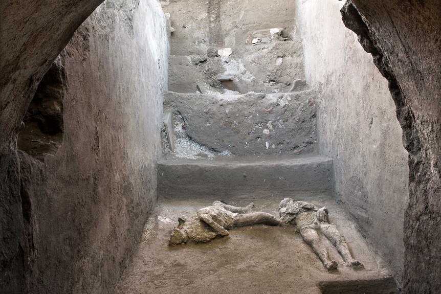 Casts of two people lay on the floor beneath stairs through which sunlight can be seen shining.