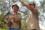 Two men armed with a gun and a knife look out towards a murky billabong.