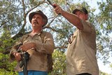 Two men armed with a gun and a knife look out towards a murky billabong.