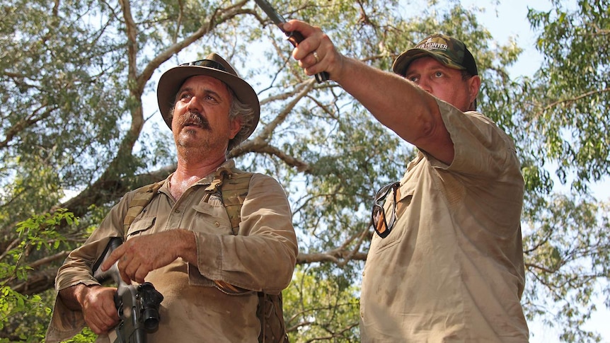 Two men armed with a gun and a knife look out towards a murky billabong.