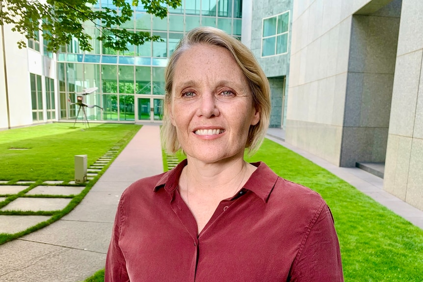 A woman with short blonde hair wearing a red blouse smiles while standing outside