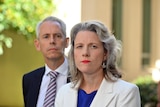 A middle-aged man and woman in suits, both with light hair, look serious in a courtyard.