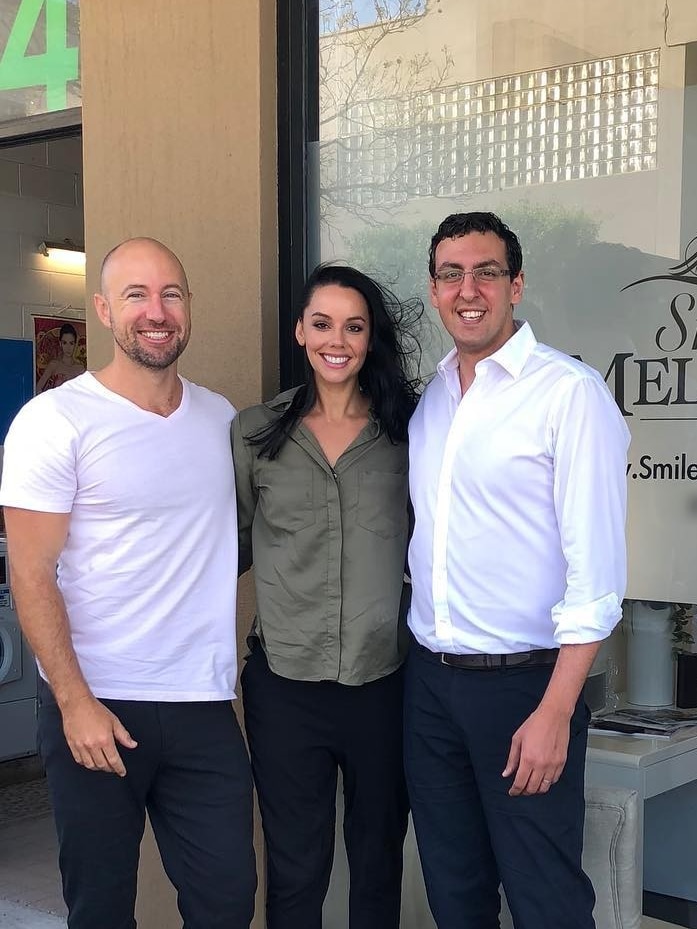 Two contestants from reality show The Block smile and pose with Ramy Georgy in front of the Smiles of Melbourne window.