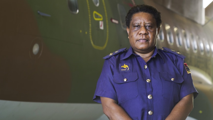 A Black woman stands next to a plane holding her arms in front of her.